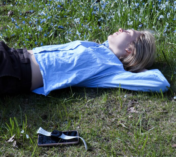 Olivia ligger på marken och njuter av solen. Smartklockan och telefonen ligger bredvid.