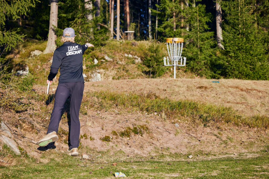 En svartklädd man kastar en frisbee mot en frisbeegolfkorg