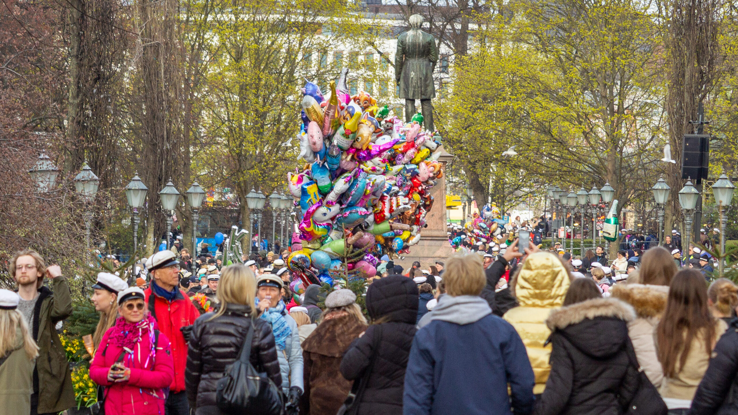 Flera människor med vita studentmössor står i Esplanadparken. I mitten av bilden syns ett stort knyppe ballonger. 